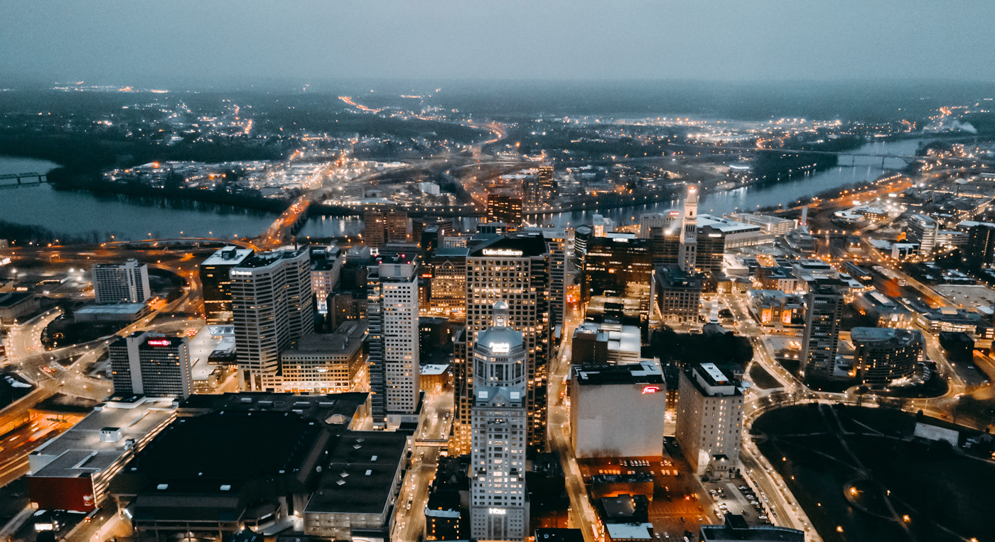 Aerial view over downtown commercial real estate CT Hartford with street lights on and some lights coming on in office buildings. The Connecticut River curves through the background under a grey sky.
