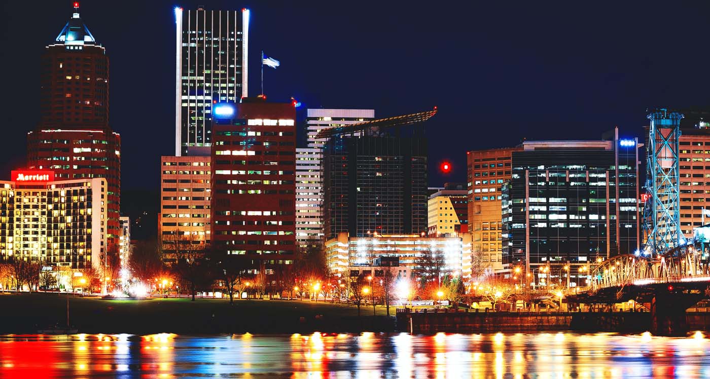 night view across the willamette river of downtown commercial real estate oregon portland office buildings with office and street lights reflecting on the river and black sky framing the buildings